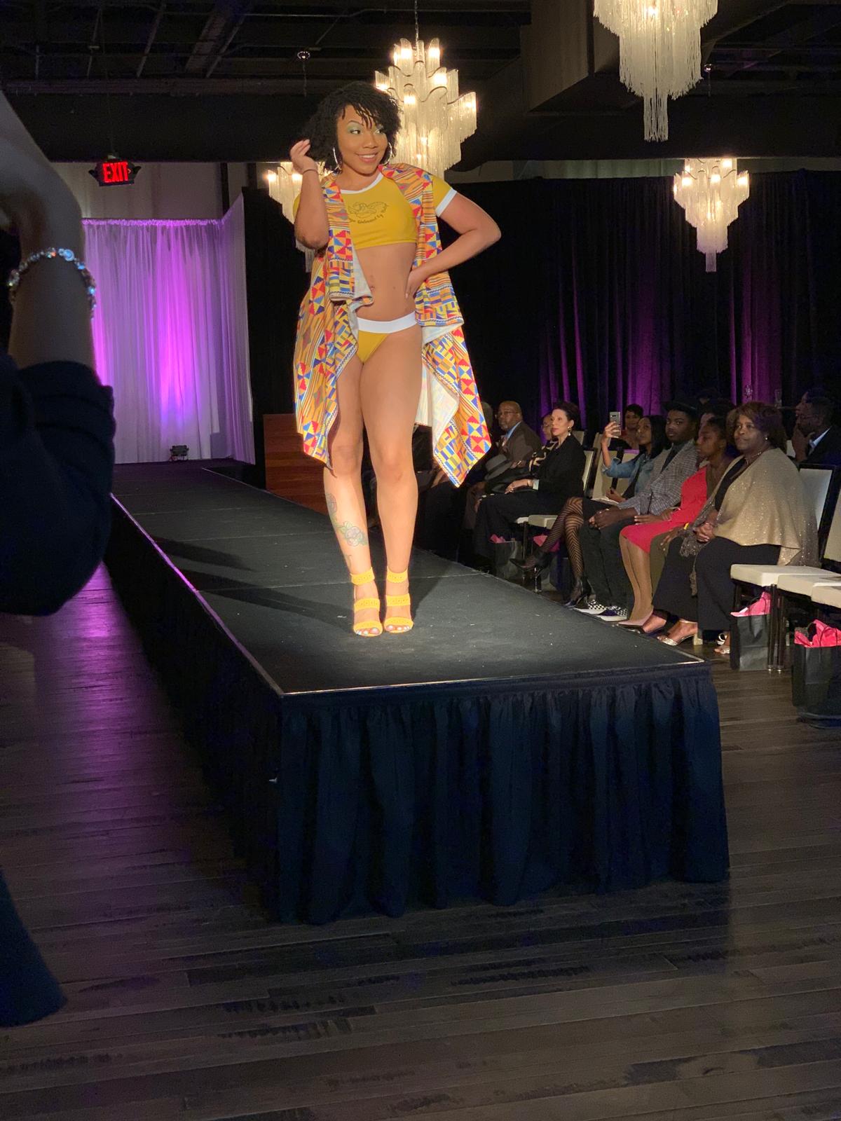 Model walking on runway in colorful outfit with audience watching, chandeliers hanging from ceiling, purple lighting in background.