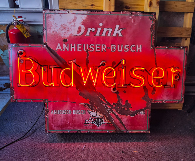Neon Budweiser sign with red background and text, mounted on a wall, partially damaged, with a fire extinguisher nearby.