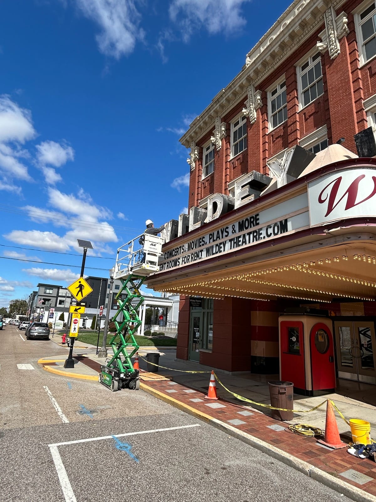Street view of a theater building with a marquee, sidewalk, and parked cars under a blue sky with clouds.