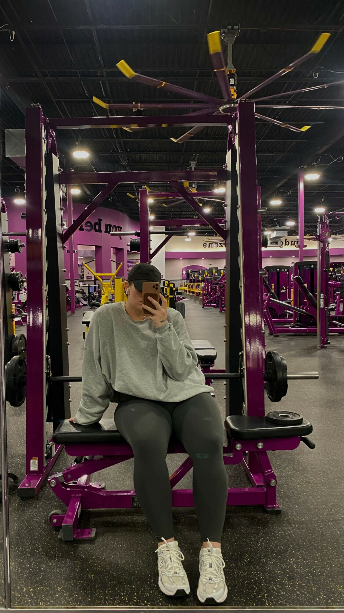 Person sitting on gym equipment in a fitness center with purple and black equipment, taking a selfie.