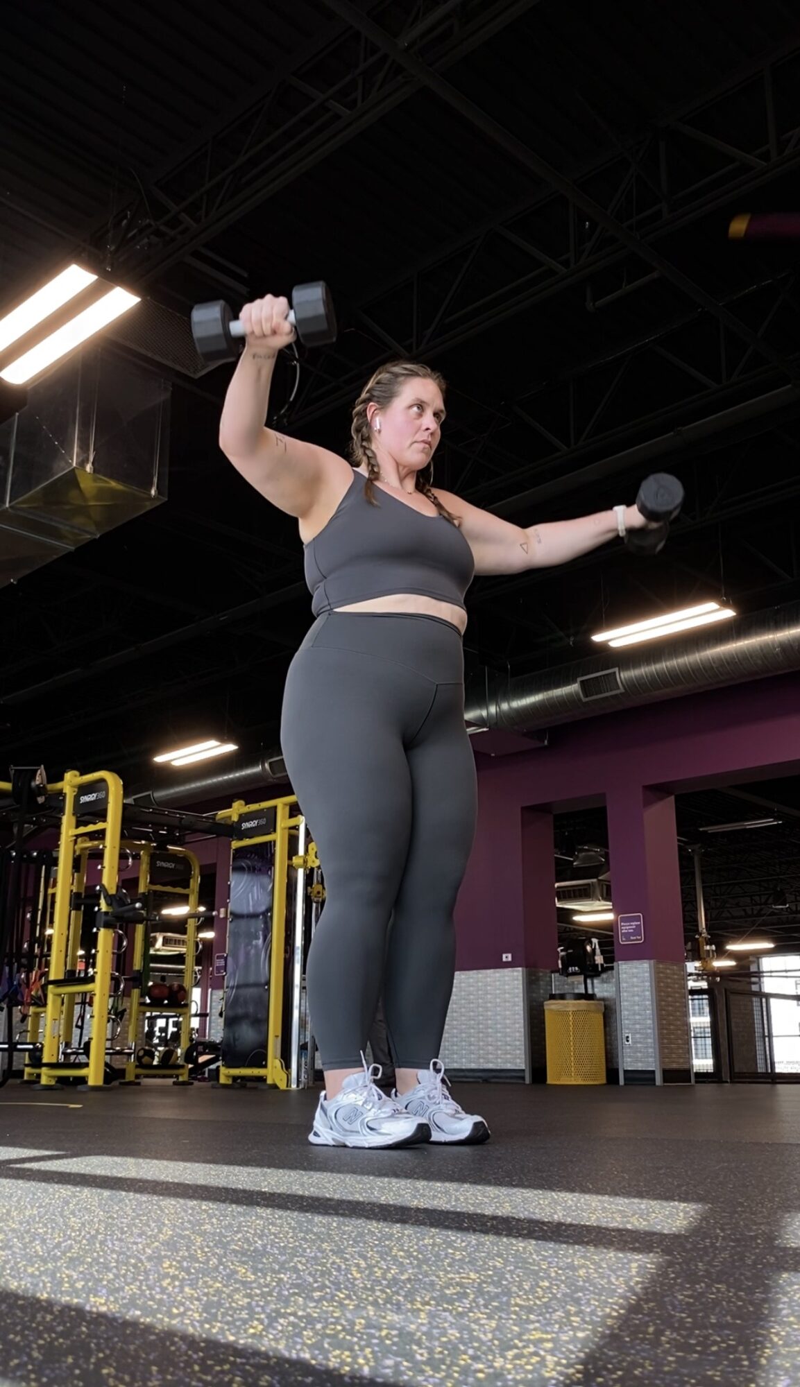 Woman lifting dumbbell in gym, holding a weight in each hand, wearing black workout clothes and white sneakers.
