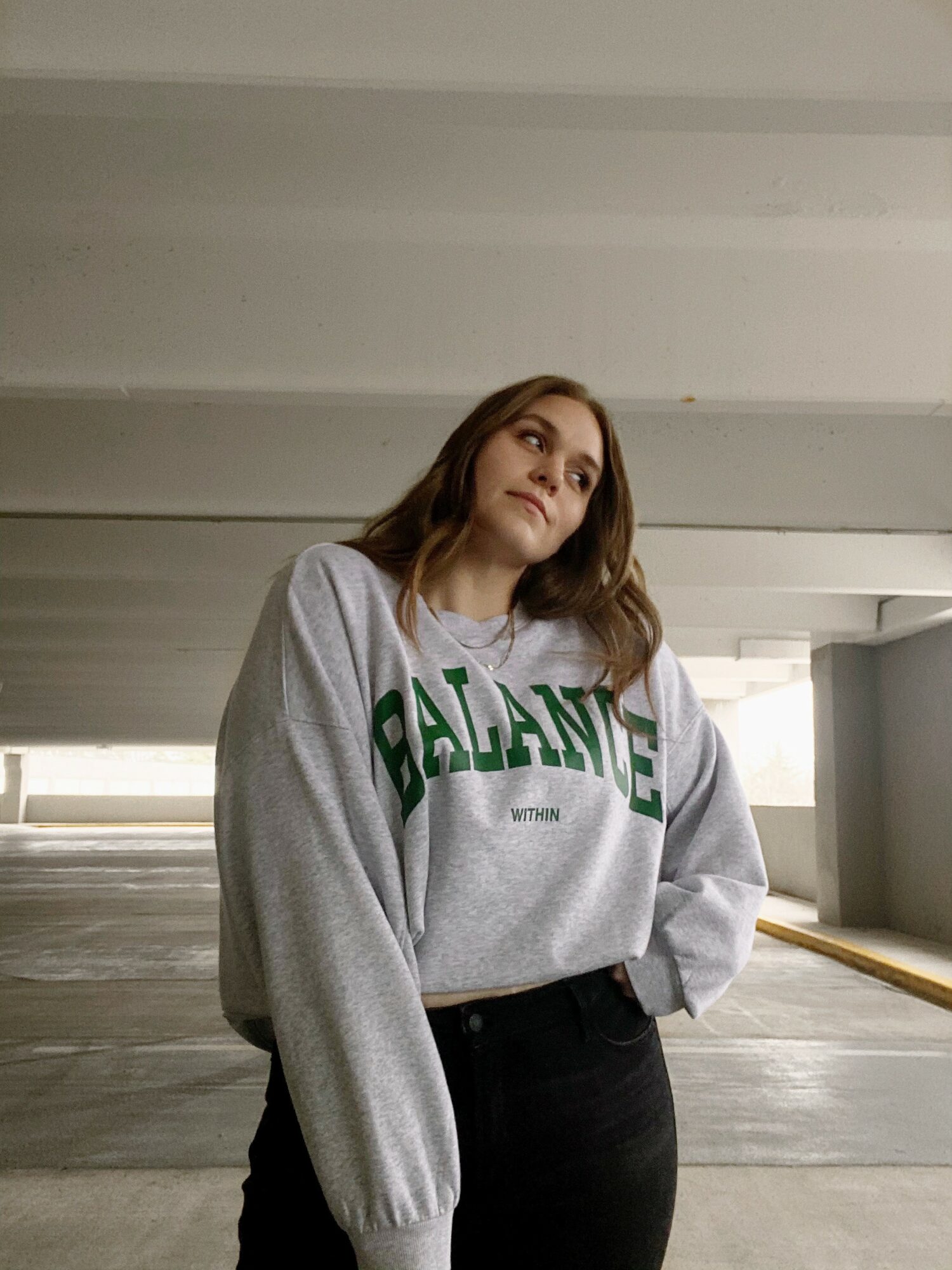 Young woman with shoulder-length hair wearing a gray sweatshirt with green text, standing in a parking garage.