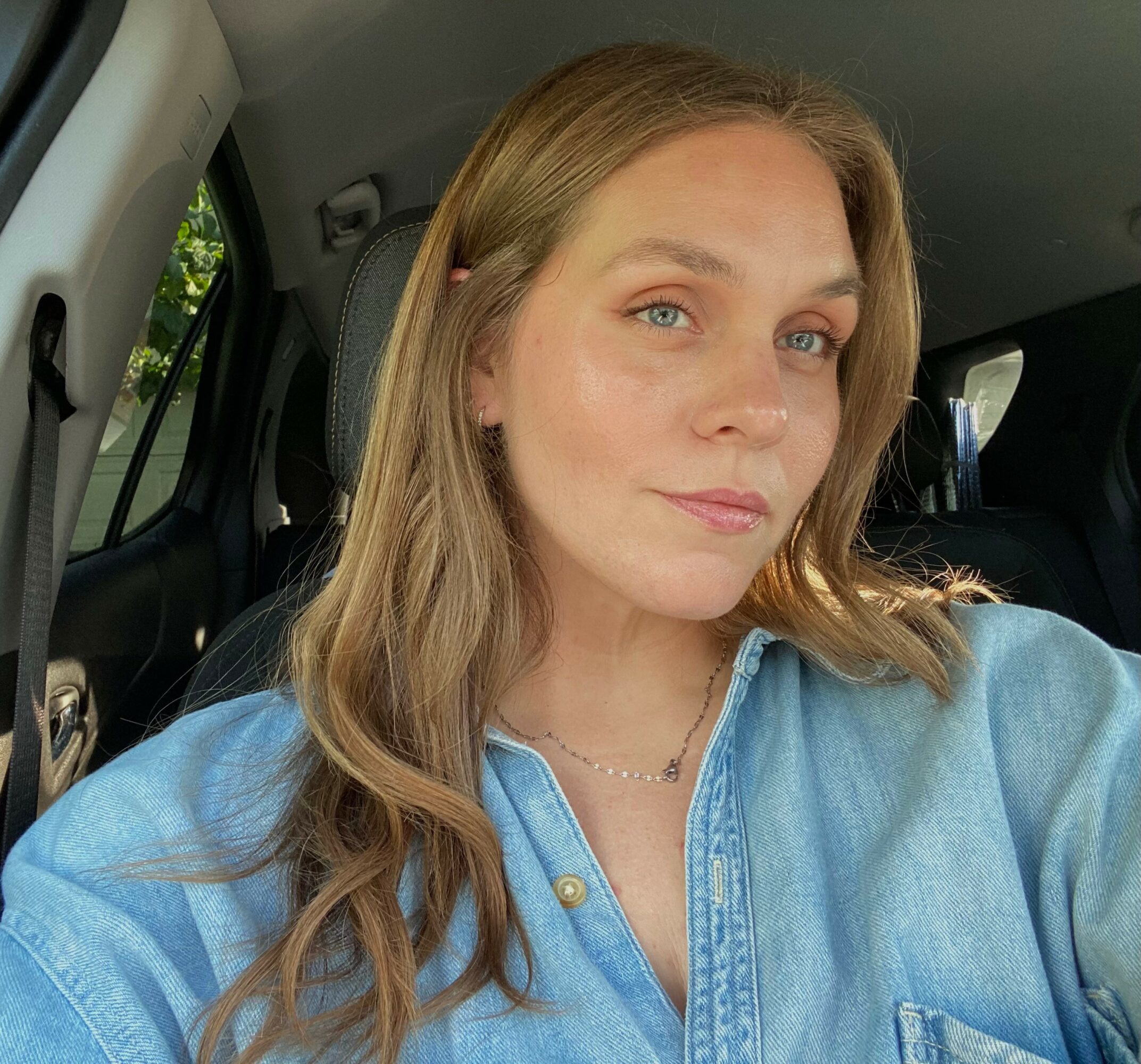 Woman with long wavy hair wearing a blue denim shirt, sitting inside a vehicle, looking at the camera.