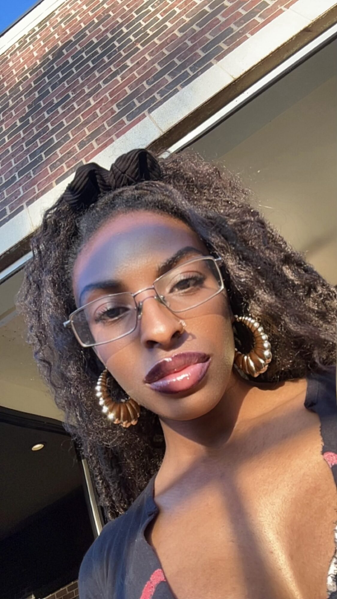 Woman with glasses and large earrings, sunlight on face, brick wall and window behind, looking at camera.