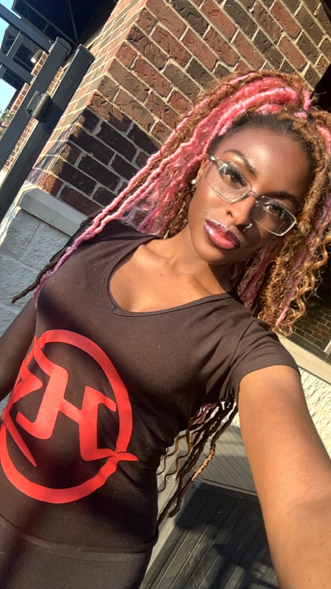 Woman with pink dreadlocks wearing glasses and a black T-shirt with a red logo, standing outdoors near a brick wall.