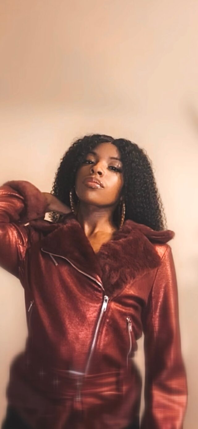 Woman with curly hair wearing a red leather jacket, posing with her hand near her head, against a plain background.