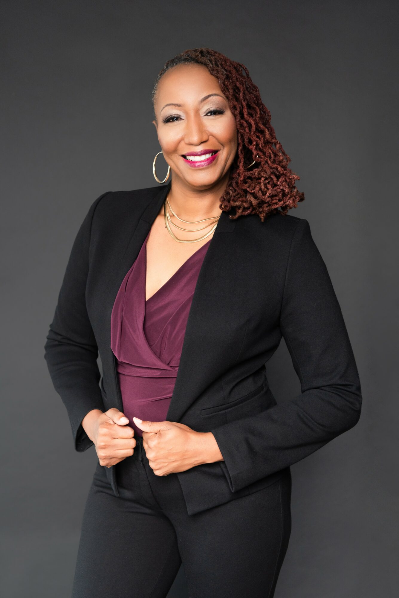 Woman with curly hair in a black blazer and maroon top, smiling, against a dark background.