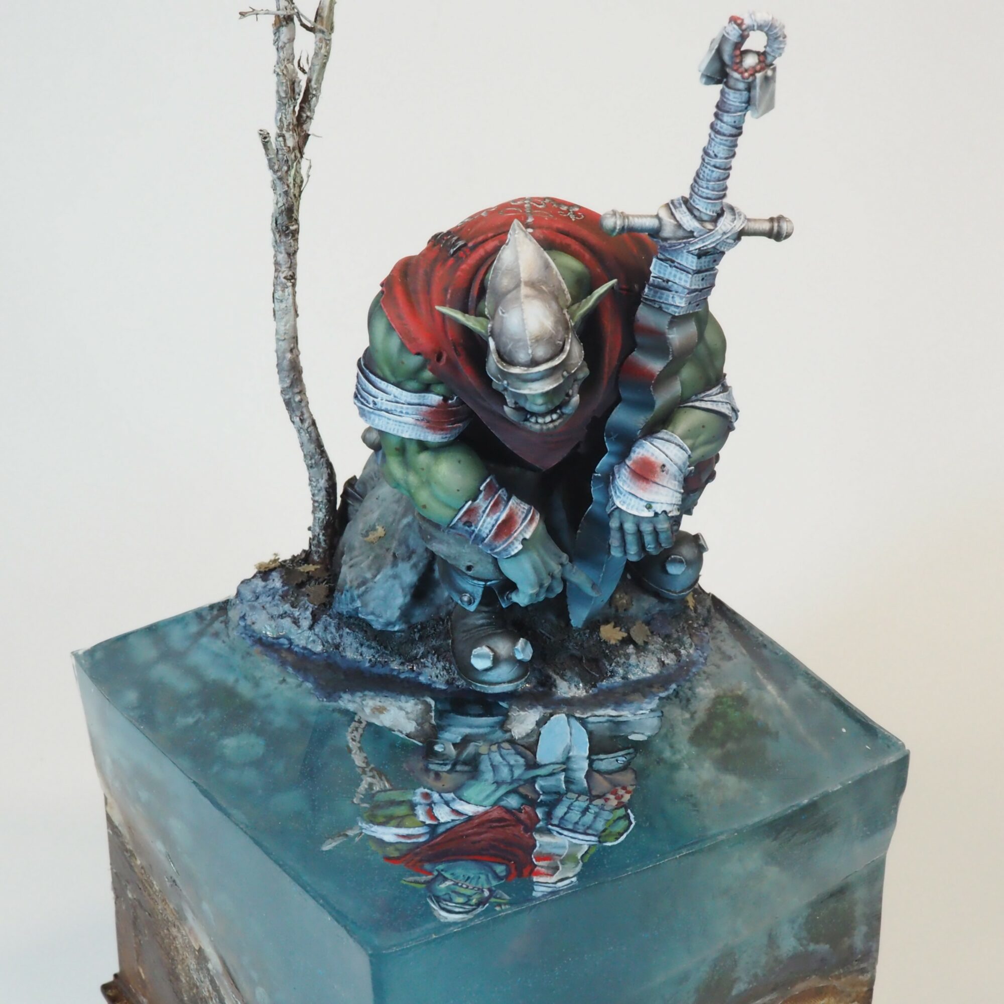 A detailed fantasy figurine of a goblin warrior with a sword, crouching on a rocky base with a tree, reflected in water.
