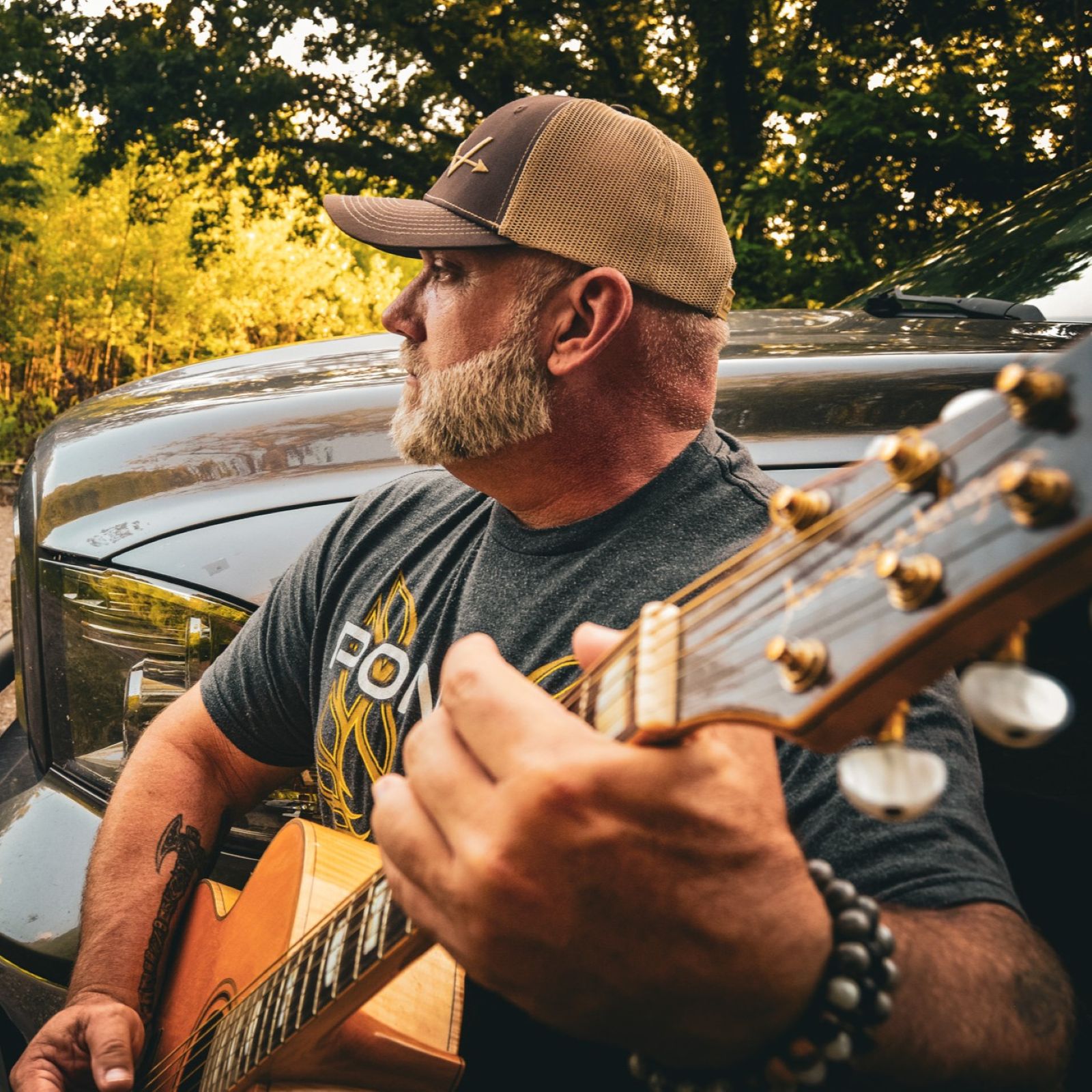 Man with a beard wearing a cap playing guitar outdoors, with trees and a vehicle in the background.