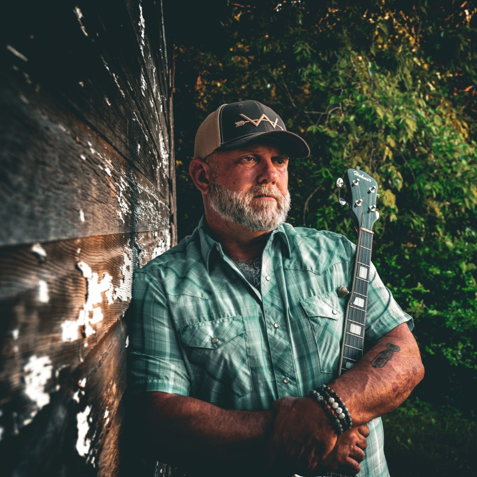 Older man with a beard and cap leaning against a dark wooden wall outdoors, surrounded by greenery.