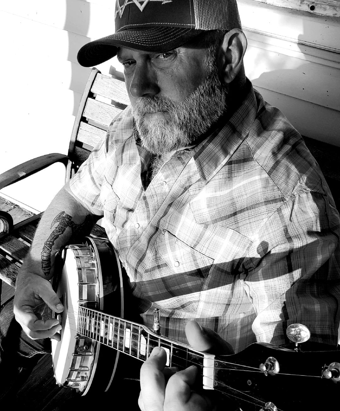 Man with a beard wearing a cap and plaid shirt playing a guitar outdoors, sitting on a bench.