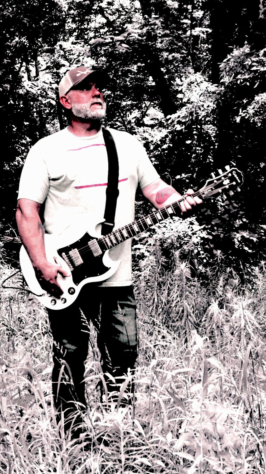 Man with sunglasses playing electric guitar outdoors among trees and tall grass.