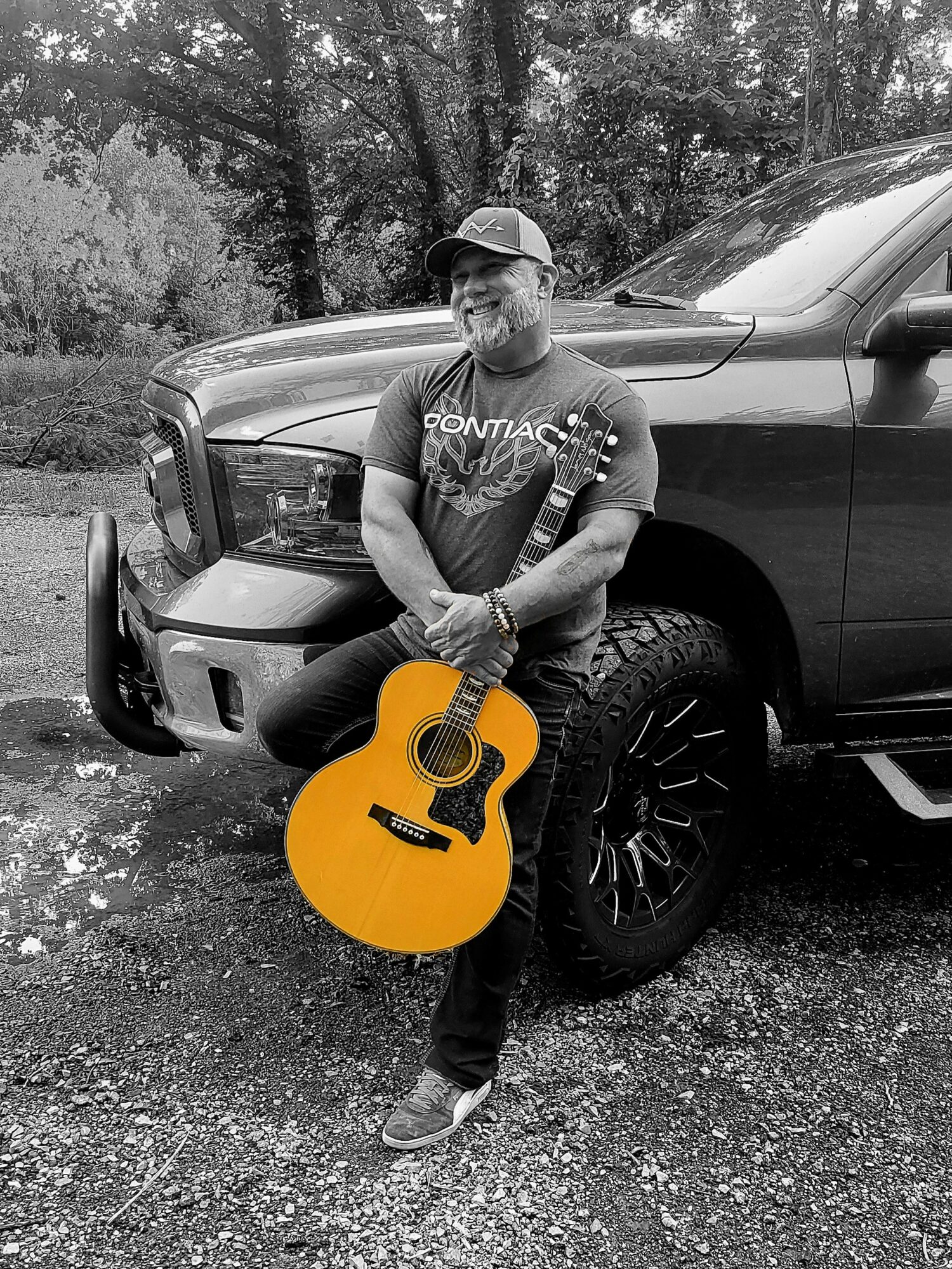 Person standing outdoors with a guitar, leaning against a vehicle, trees in the background, black and white image.