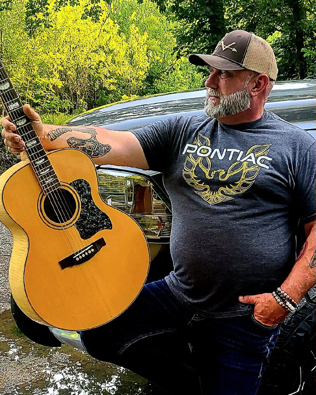 Man with a beard and cap holding an acoustic guitar outdoors, trees and a vehicle in background.