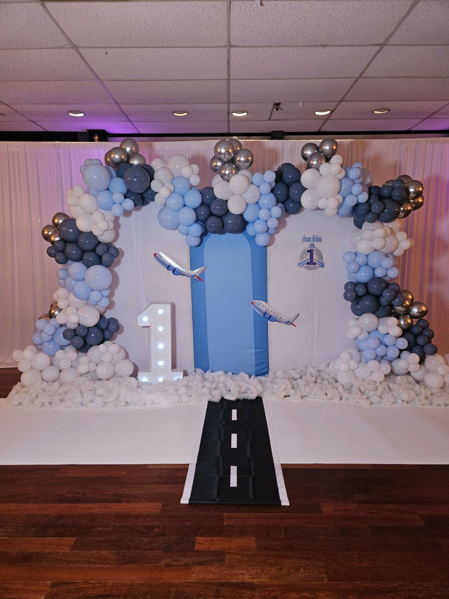 Party decoration with balloons in blue, white, and silver, and a blue gift box on a decorated backdrop.