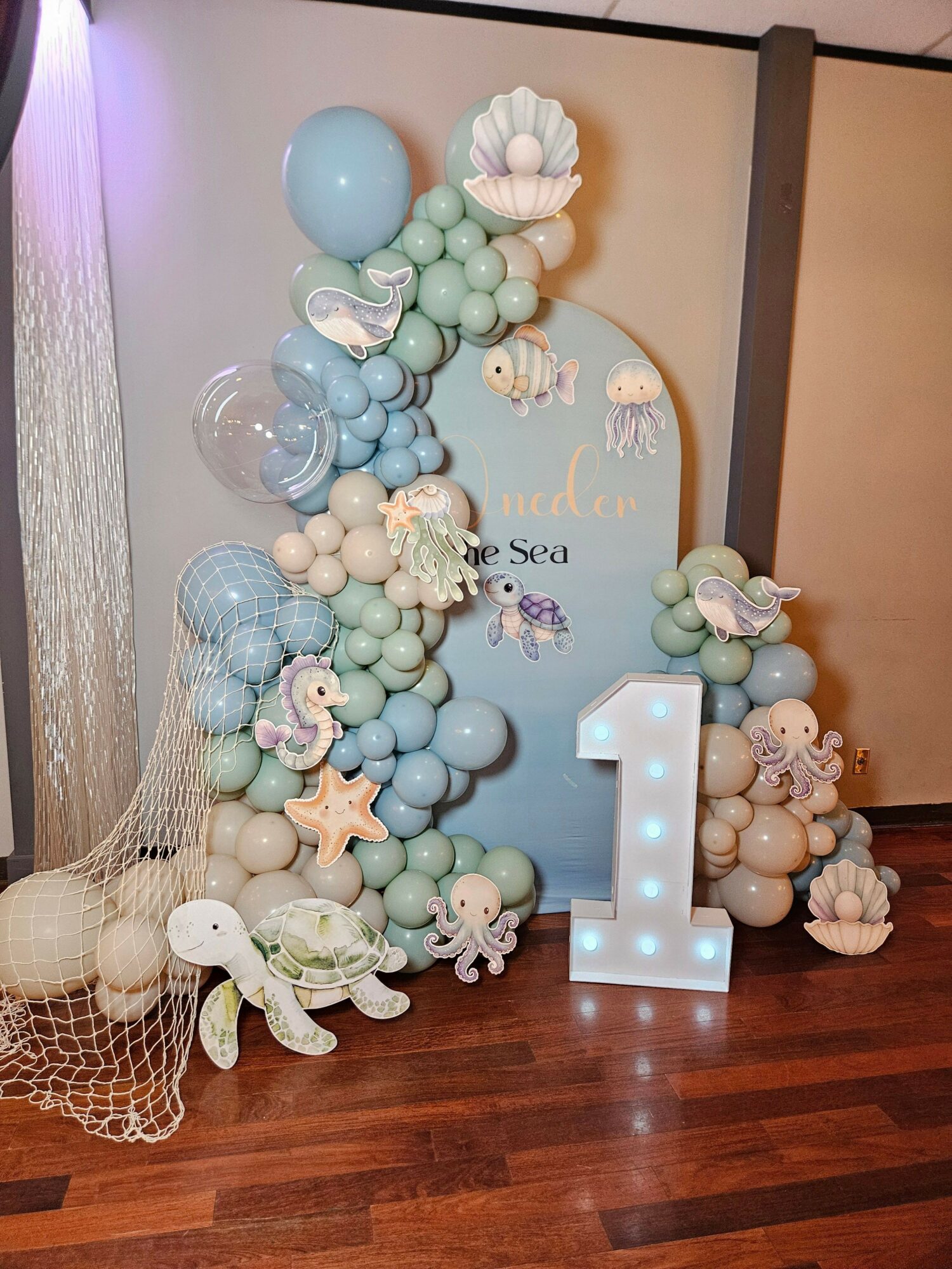 Decorative arrangement of pastel-colored balls, seashells, and a large illuminated letter 'L' on a wooden floor.
