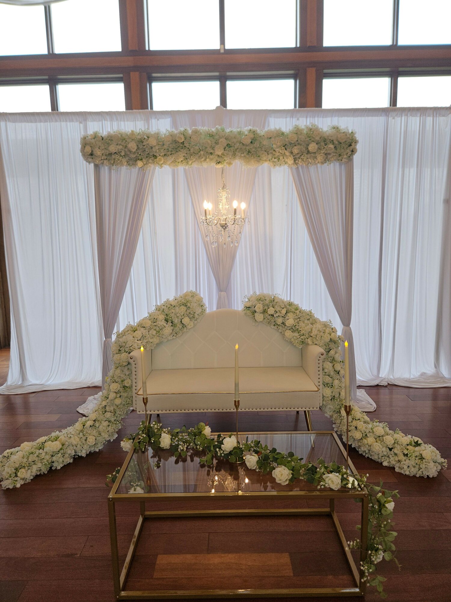 Wedding ceremony setup with a decorated arch, white curtains, and floral arrangements on a wooden floor.