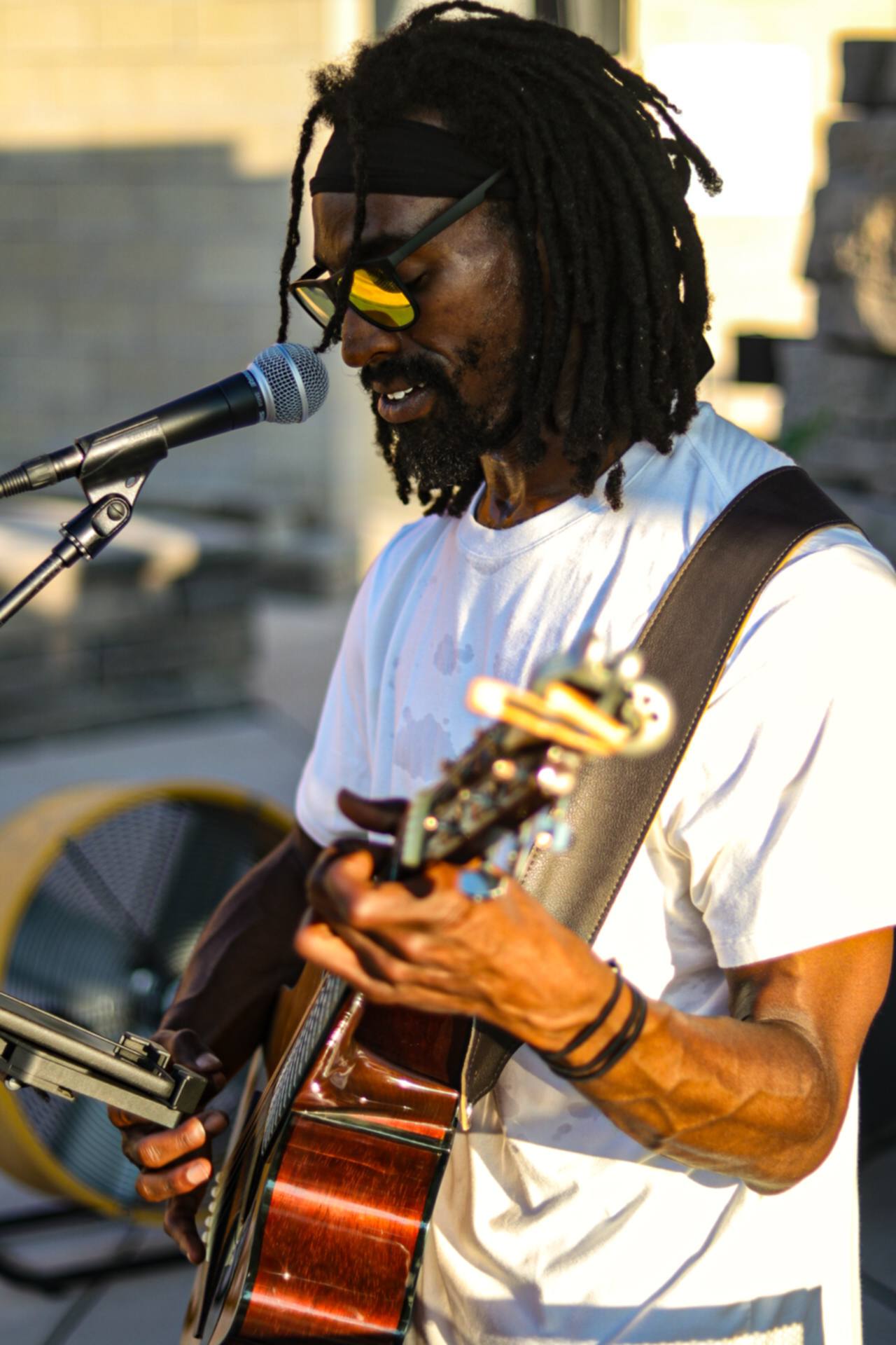 Musician with dreadlocks, sunglasses, and white t-shirt playing guitar and singing into a microphone outdoors.