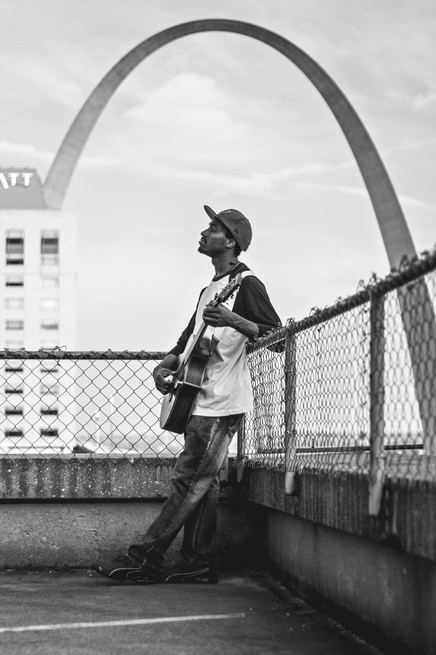 Person standing on a rooftop with a guitar, wearing a hat and sunglasses, with the Gateway Arch in the background.
