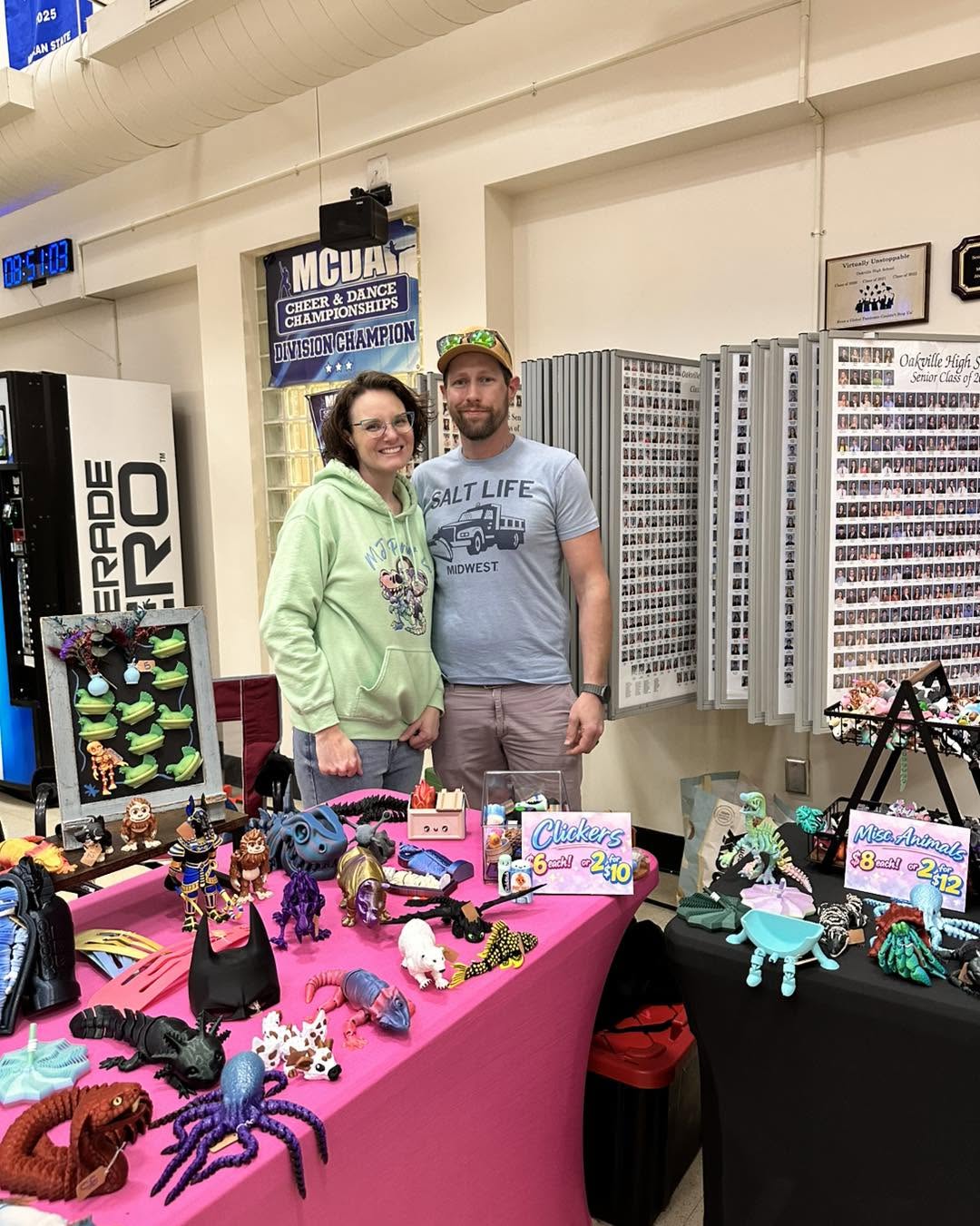 Two people stand behind a table with colorful crafts and decorations at an indoor event, smiling at the camera.