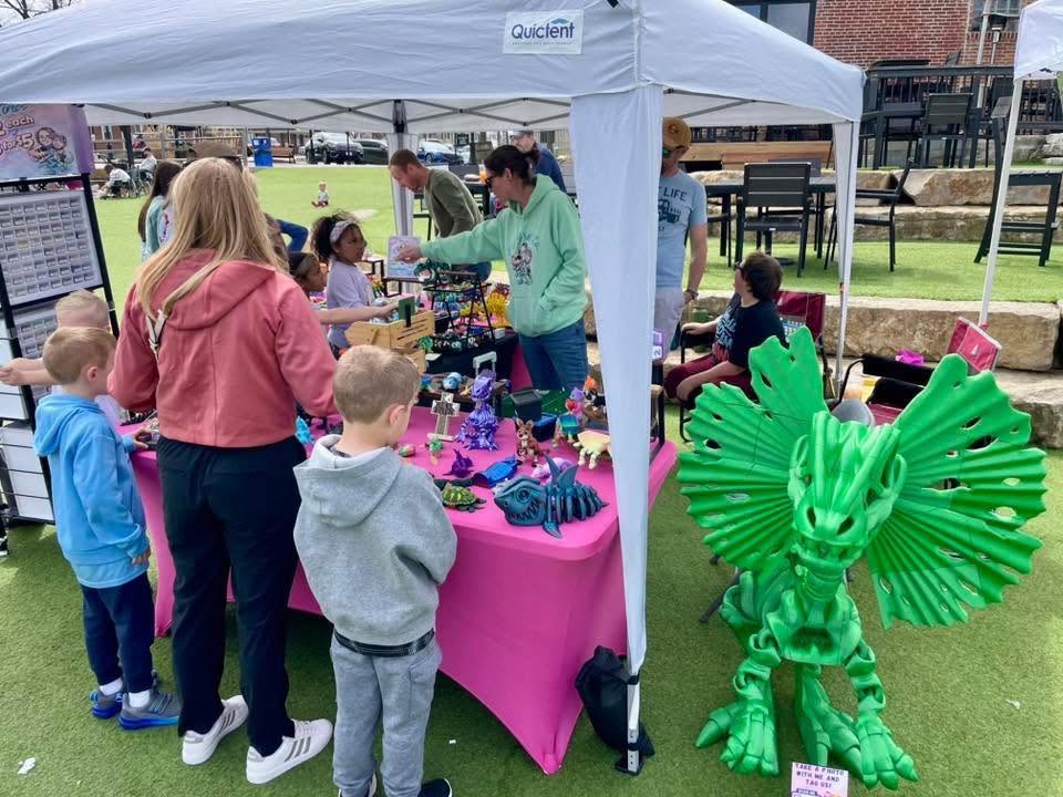Children and adults gather around a pink table under a white canopy at an outdoor event, with a green dragon sculpture nearby.