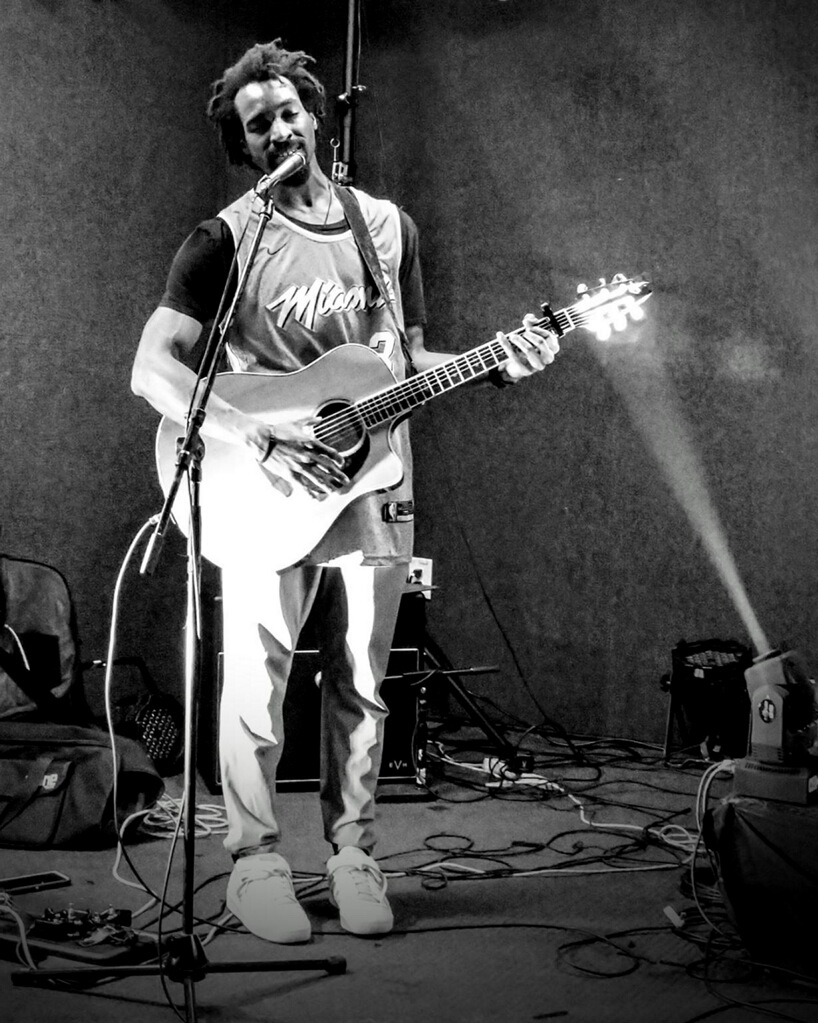 Person playing guitar and singing into microphone on stage, with equipment and cables around, in black and white.
