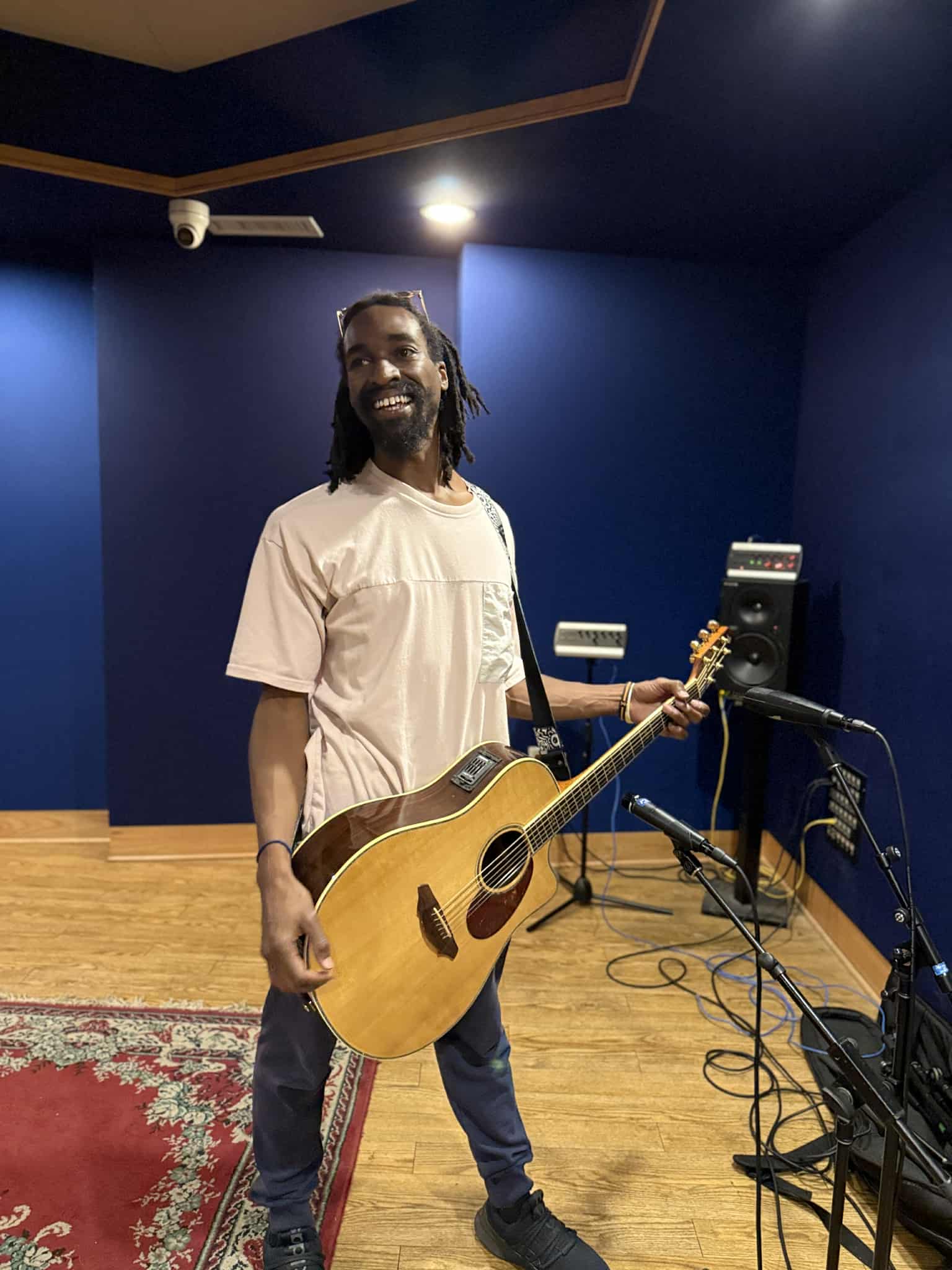Person holding an acoustic guitar in a music studio with blue walls and equipment, smiling and looking at the camera.