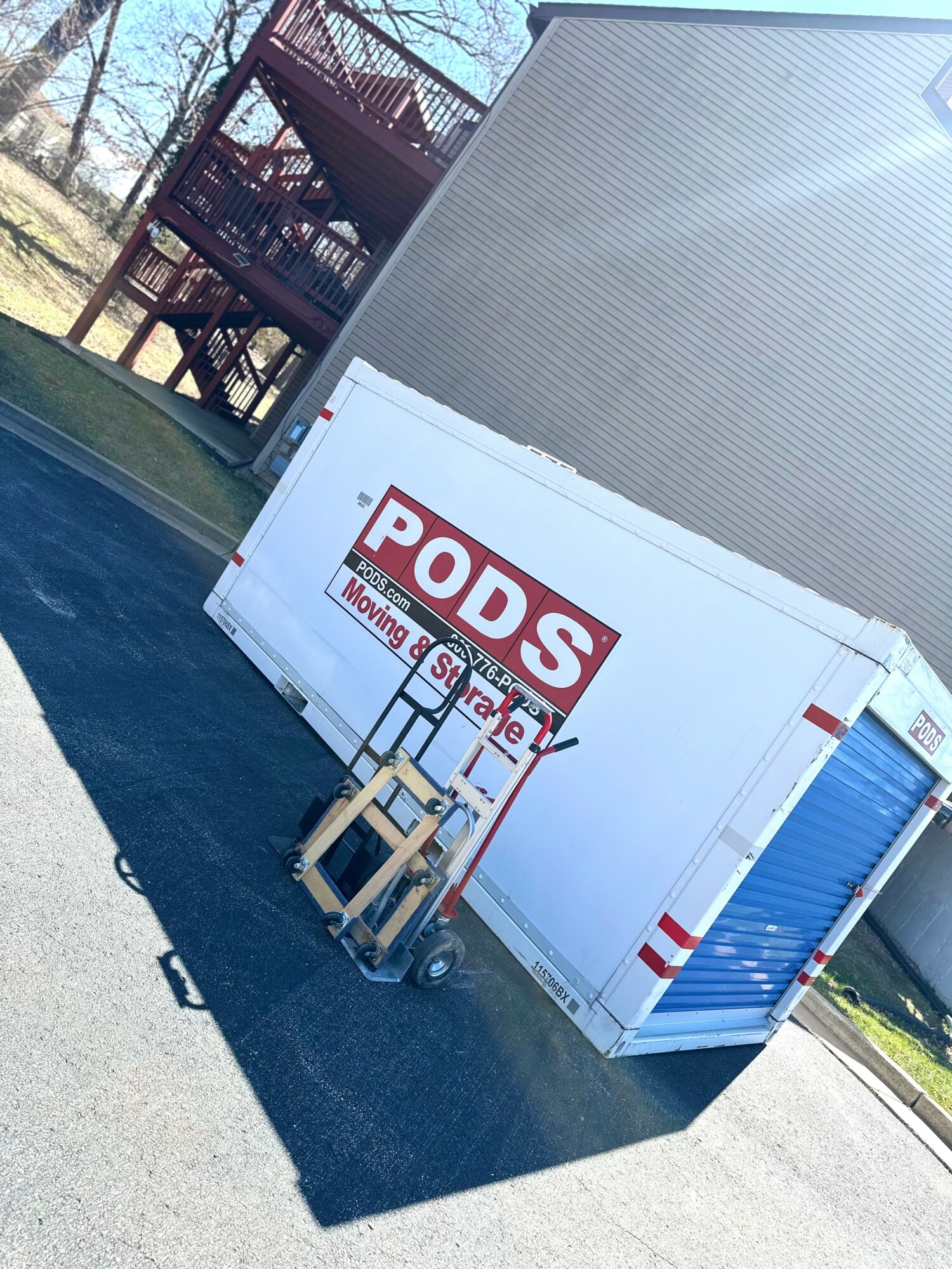 A large white PODS storage container with a red and white sign, parked on a paved surface near a building and a wooden staircase.