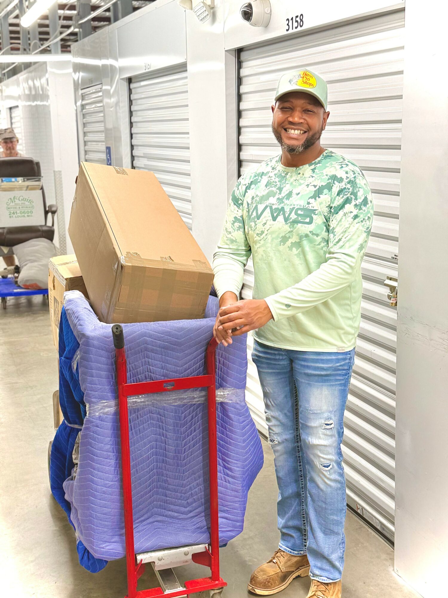 Smiling man in a warehouse, wearing a cap and casual clothes, standing next to a cart with a large box and blue packing material.
