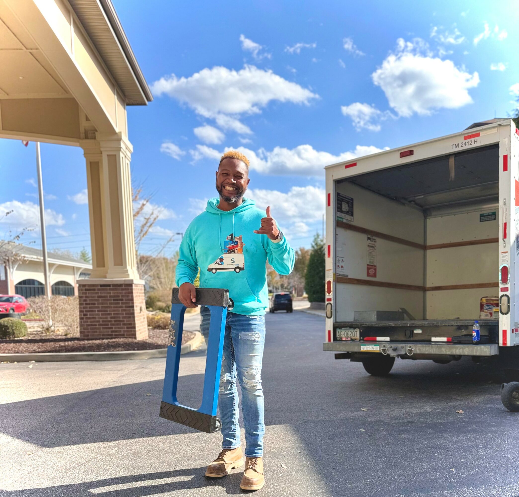 Man standing outdoors holding a framed picture, smiling, with a delivery truck and building in background, under a blue sky with clouds.
