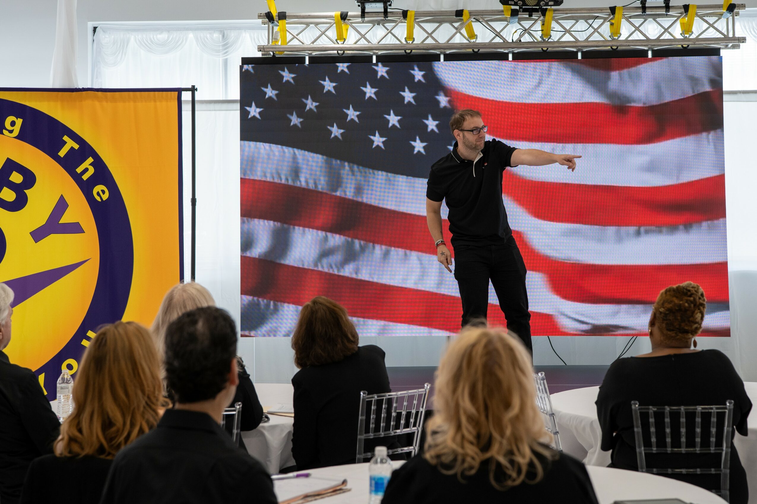 Person on stage pointing at audience with American flag backdrop, audience seated facing stage, some with blonde hair, some with dark hair.