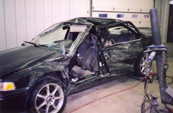Damaged black car with a shattered windshield and deployed airbags inside a garage.