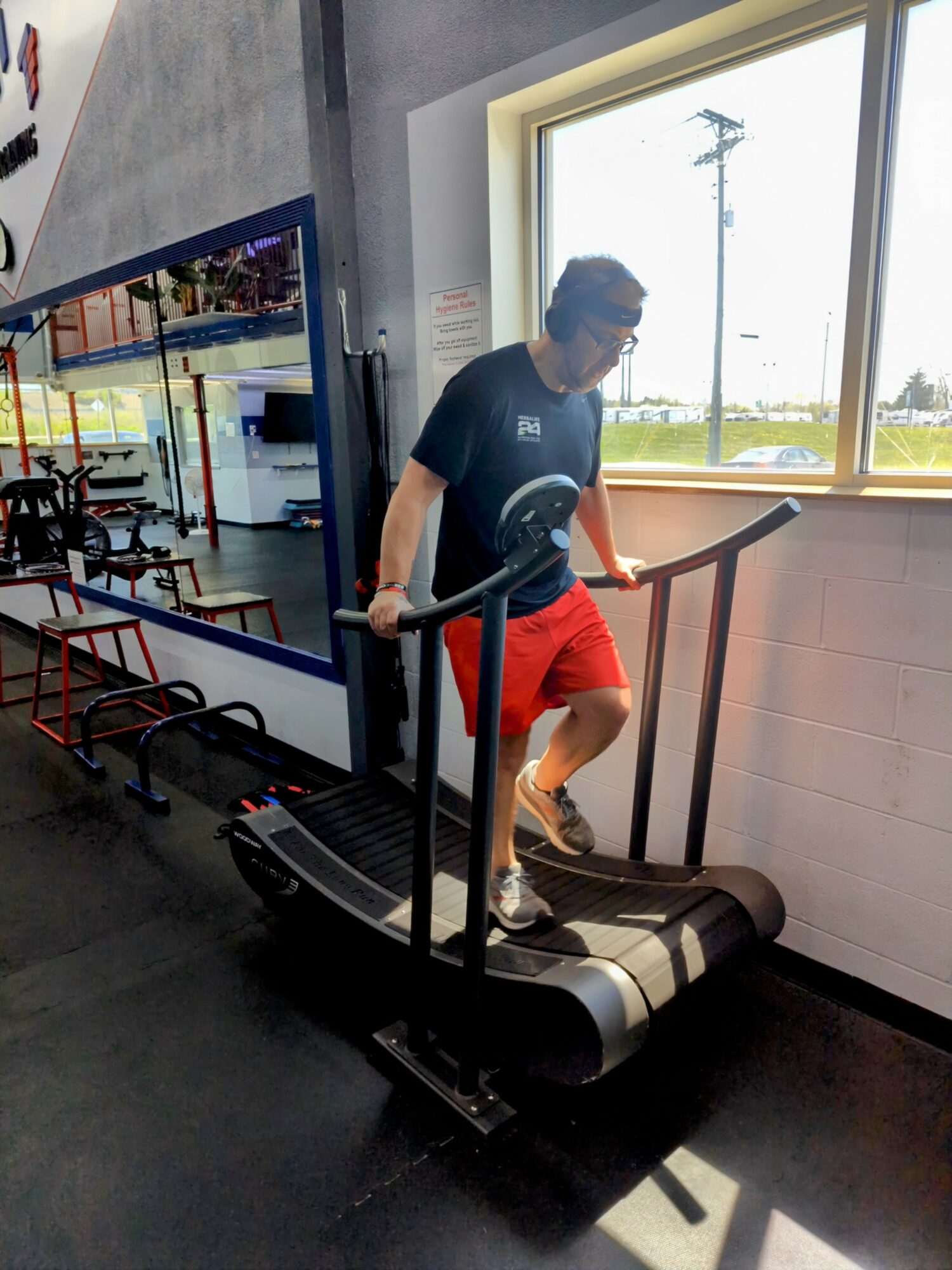 Person walking on a treadmill in a gym, wearing a black shirt and red shorts, near a large window.