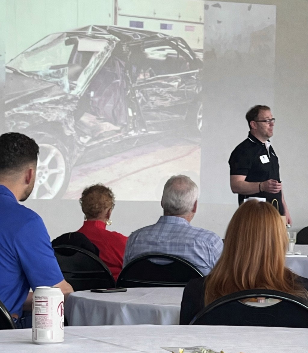 A presentation shows a damaged car on a screen, with a speaker and an audience seated at tables.