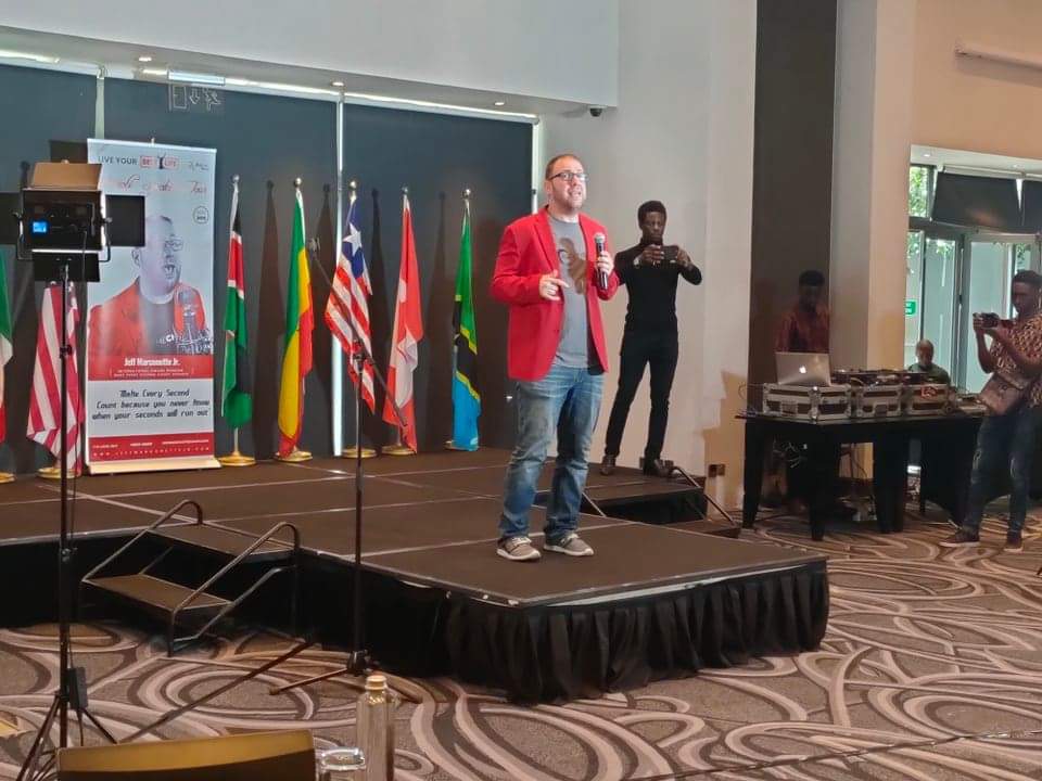 Man in red blazer speaking on stage with flags behind him, audience and event staff nearby.