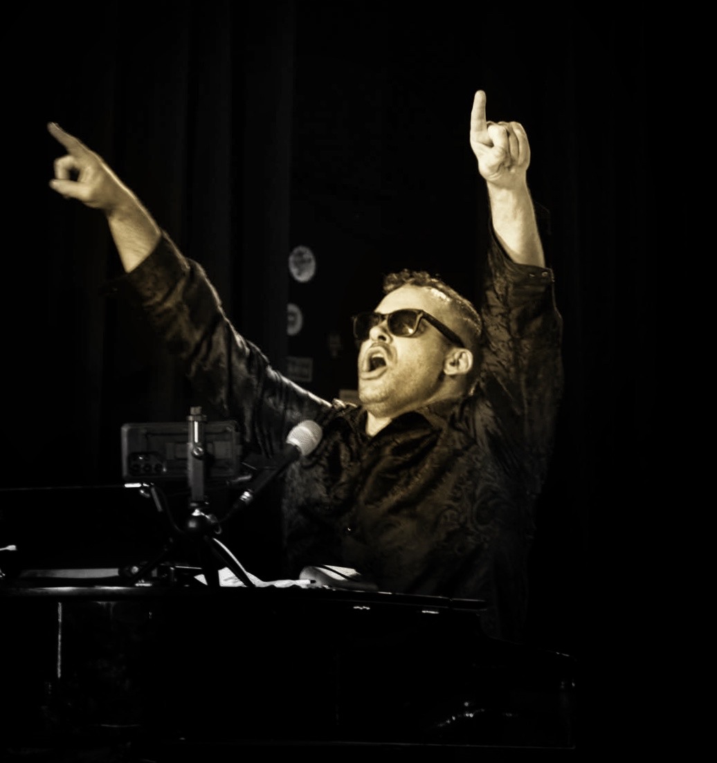 Person wearing sunglasses singing or speaking with arms raised, on stage with a microphone and dark background.