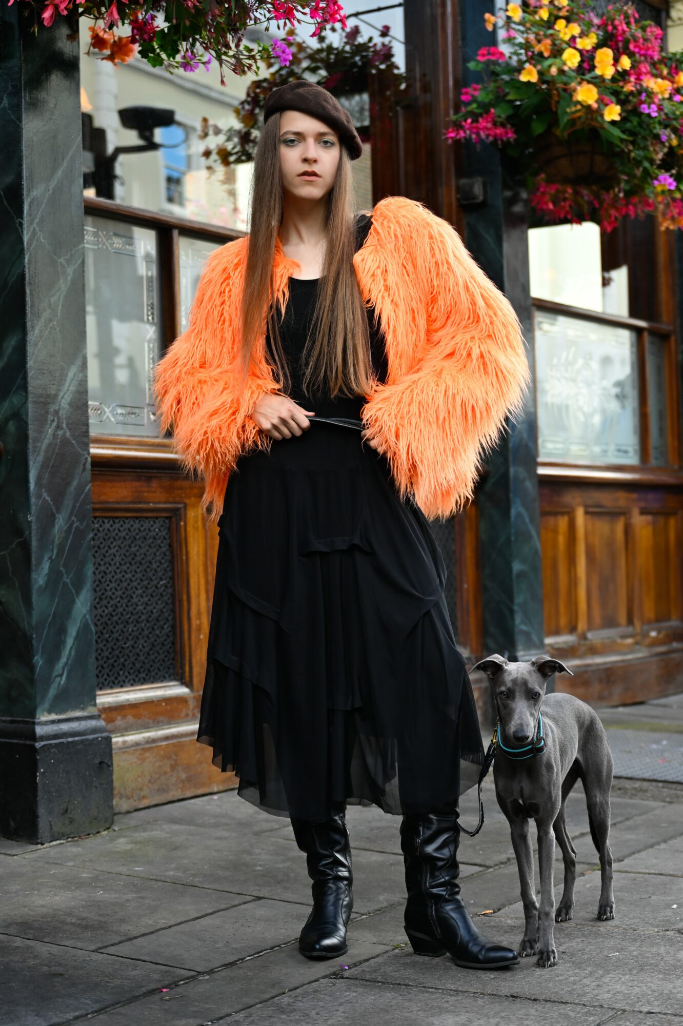 Young woman with long hair in black dress and orange furry jacket standing on sidewalk with dog, colorful flowers in background.