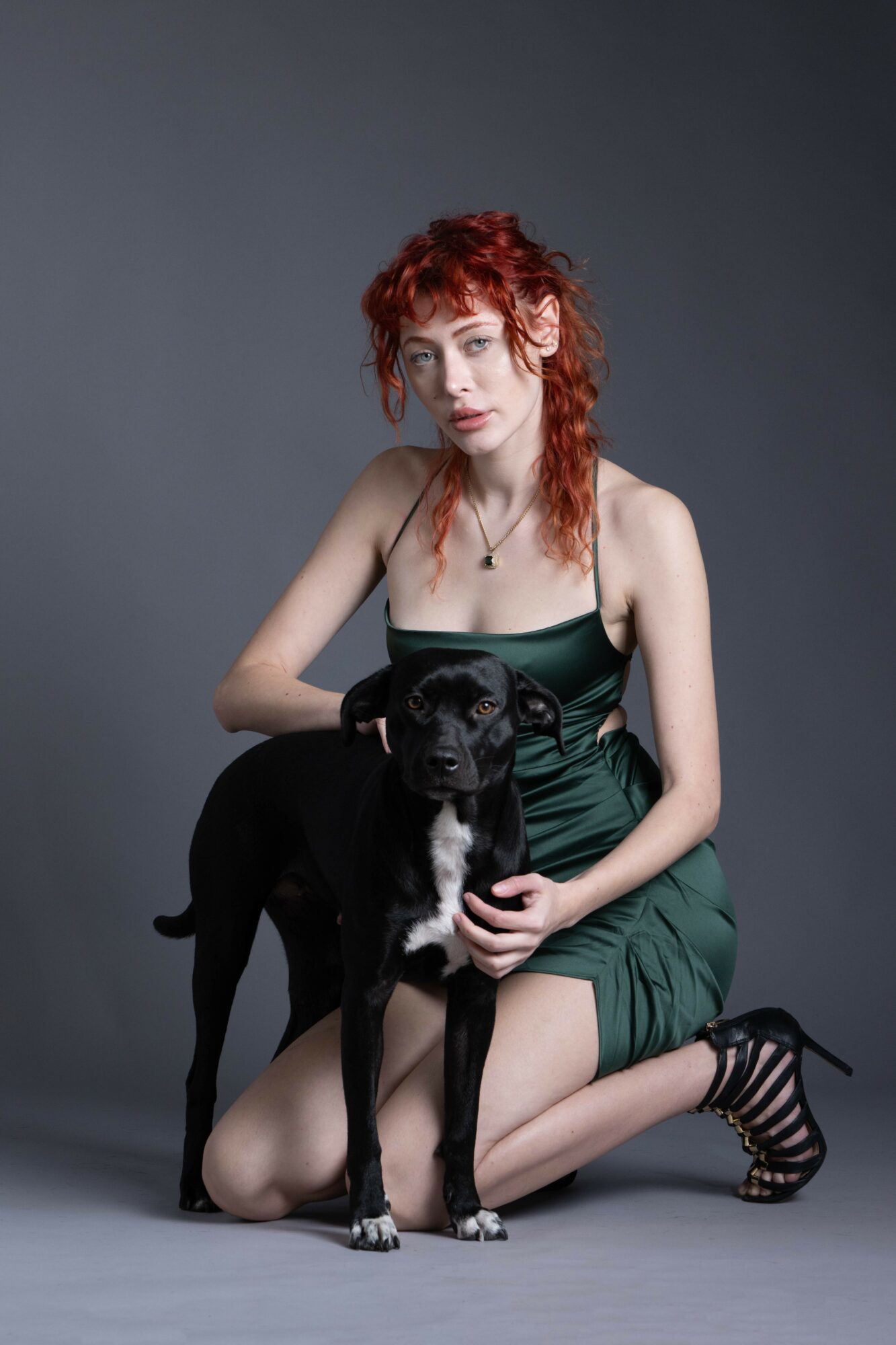 Young woman with curly red hair kneels beside a black dog against a gray background.