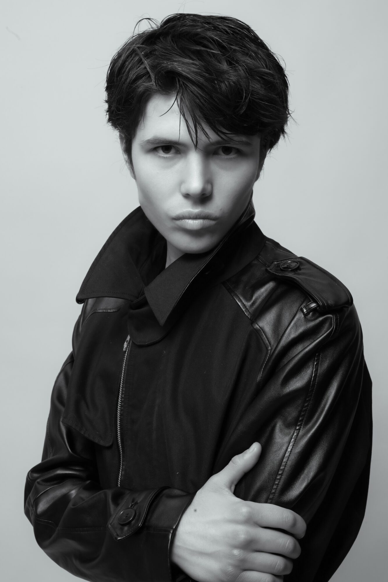 Young person with short dark hair wearing a black leather jacket, looking directly at the camera with a serious expression.