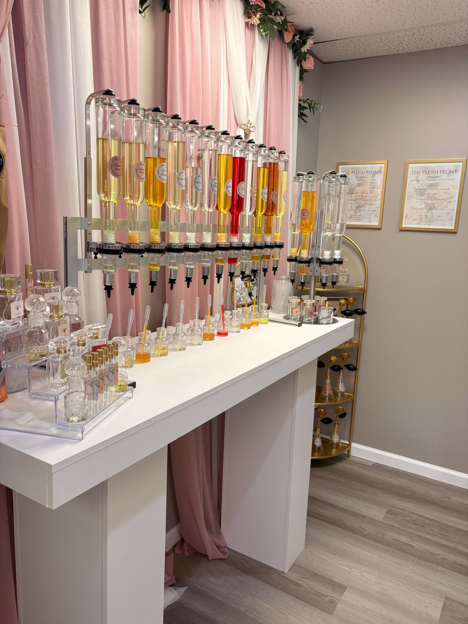 Display of various lip glosses and beauty products on a white table with pink curtains in the background.