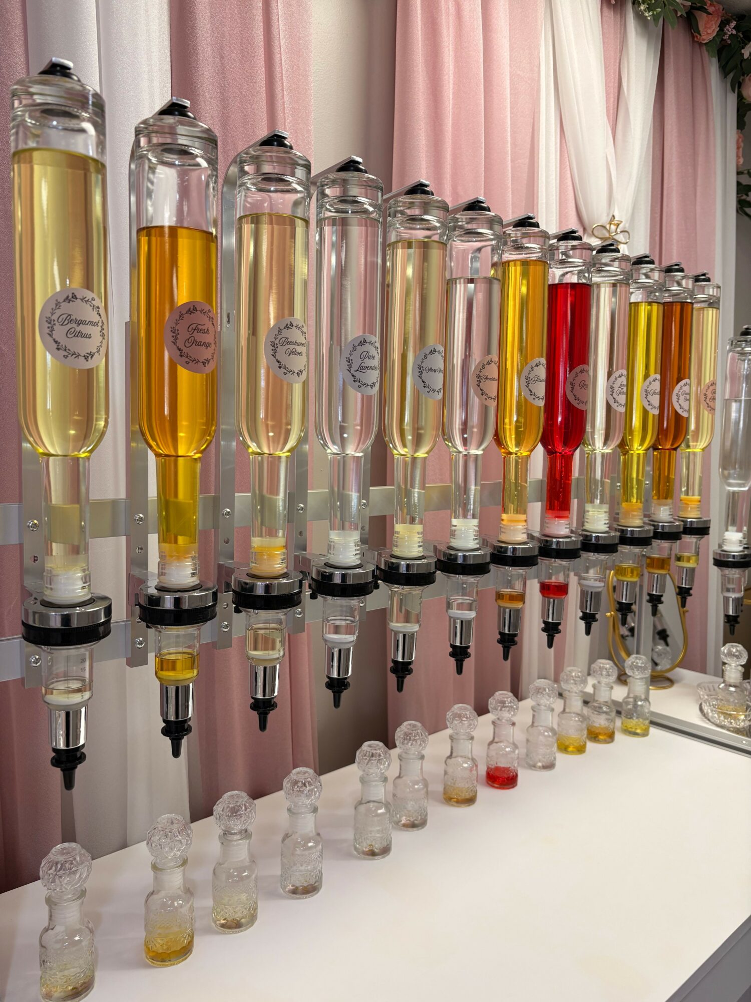 Multiple glass bottles with yellow liquid and droppers, arranged on a white surface with pink curtains in background.