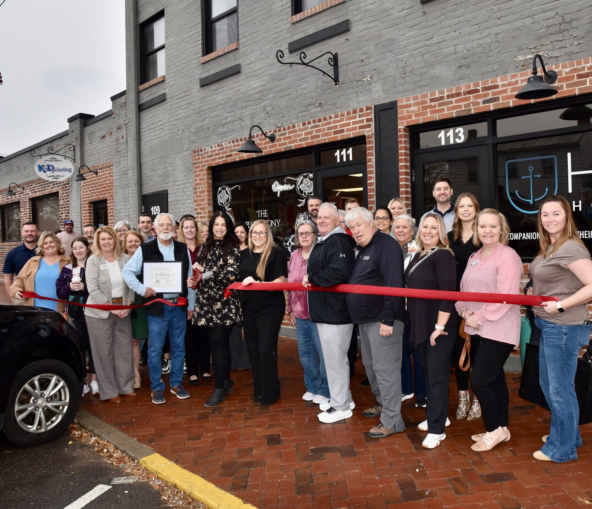 Group of people gathered outside a building for a ribbon-cutting ceremony, with a red ribbon in front of the group.