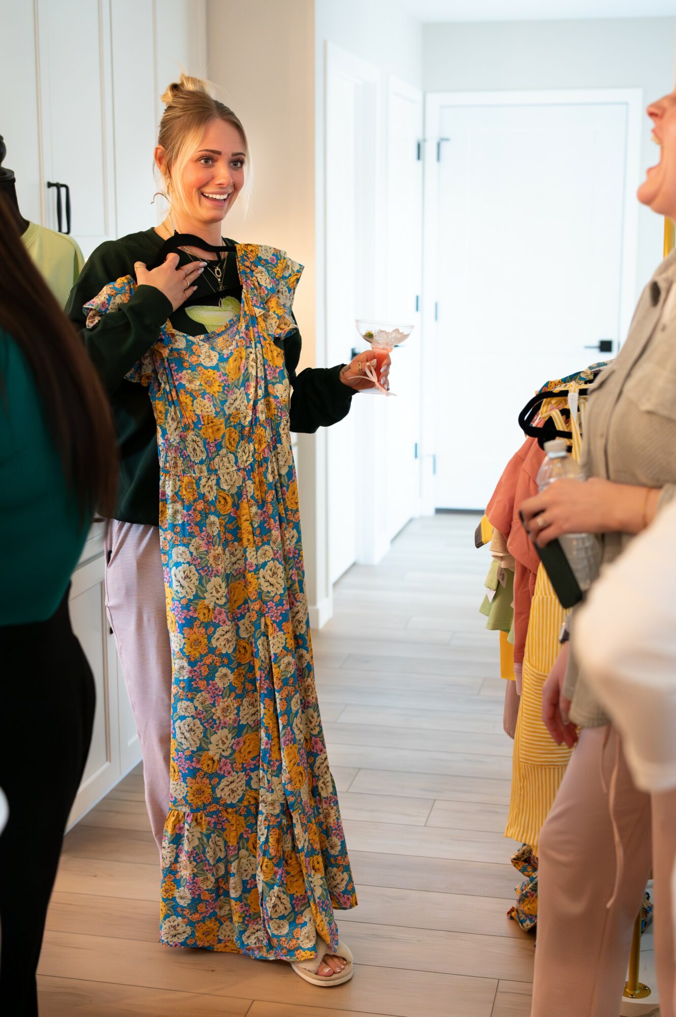 Group of women talking indoors, one woman holding a glass, smiling, wearing a long floral dress, bright room with white walls.