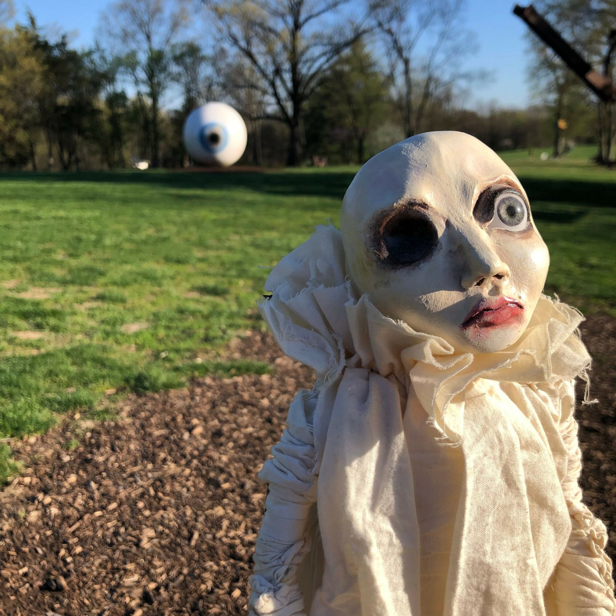 Person wearing a realistic animal mask with large eyes and open mouth, outdoors in a park with trees and grass.
