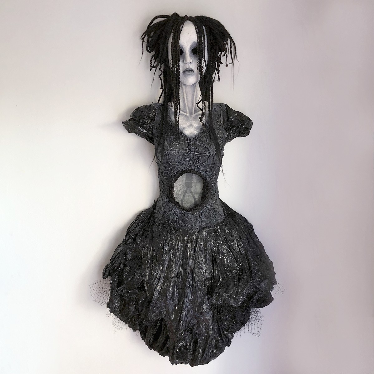 A doll with a gothic appearance, black dress, and dreadlocks, mounted on a wall against a plain background.