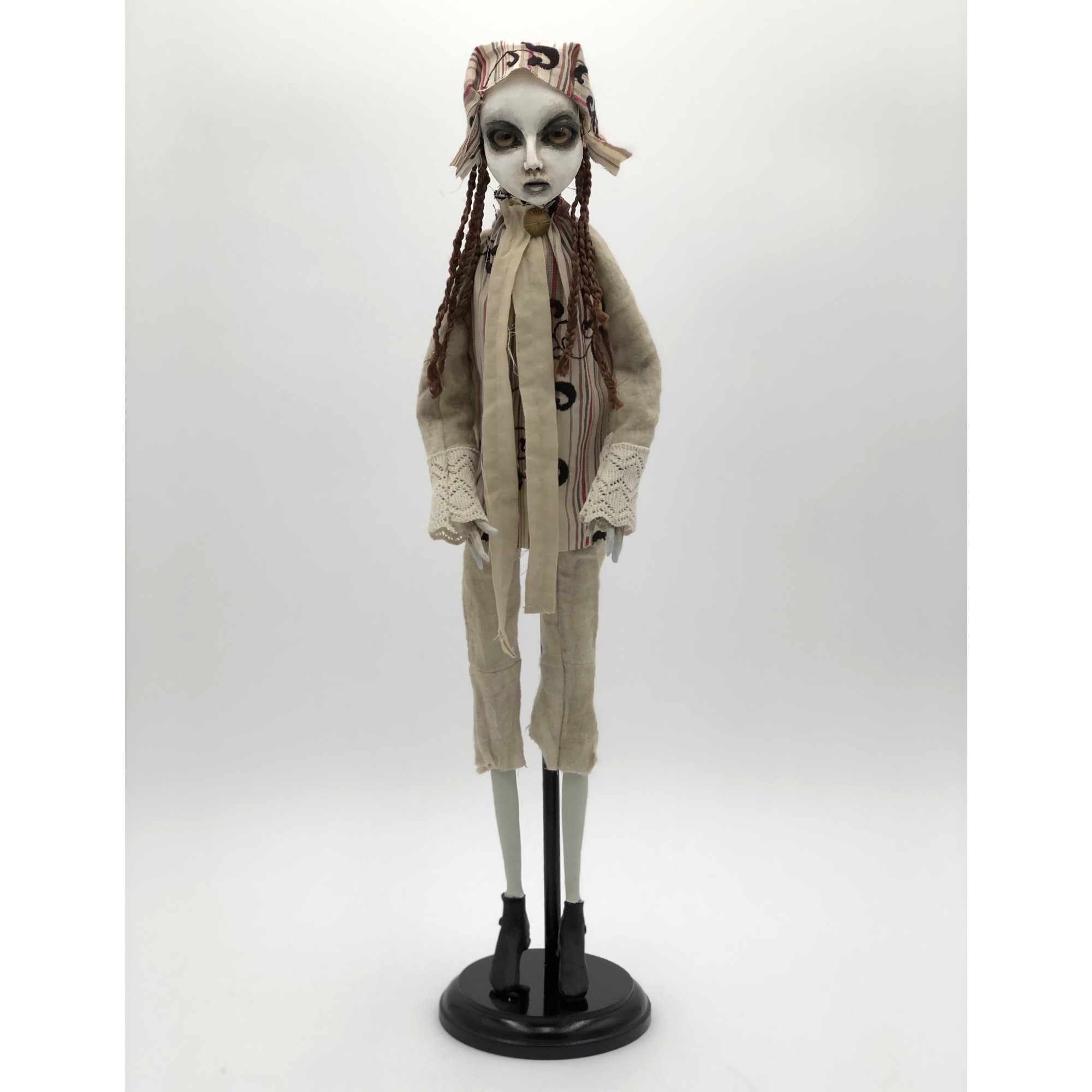 A doll-like figure with long limbs, wearing a beige outfit, with detailed makeup and braided hair, standing on a black base.