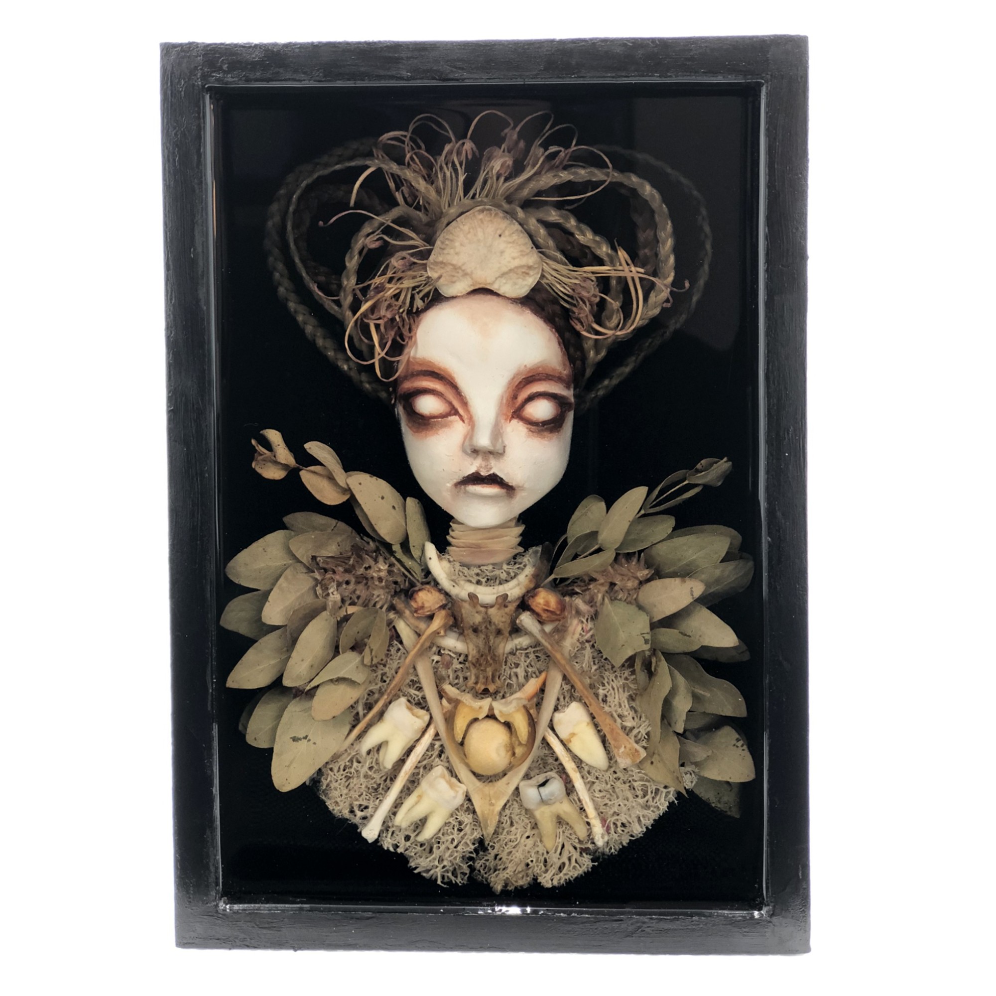 A doll with a pale face, dark makeup, and elaborate hair, dressed in a costume with leaves and lace, inside a black box.
