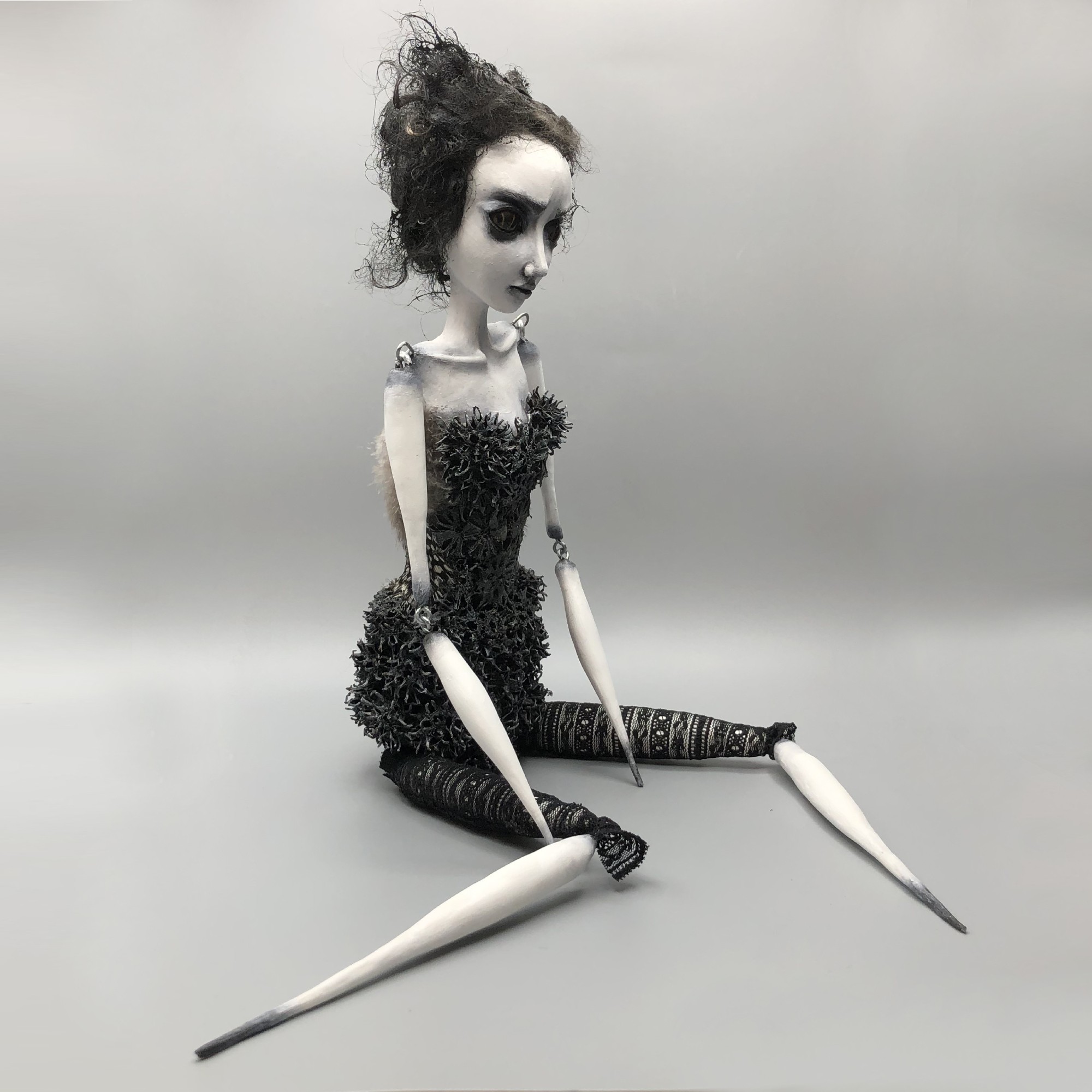 Doll with dark curly hair, sitting on the ground, wearing a textured black dress and patterned leggings, with a neutral background.