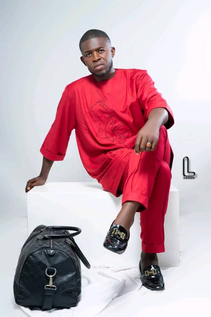 Young man in red outfit sitting on a white block, with a black bag on the floor, against a plain background.
