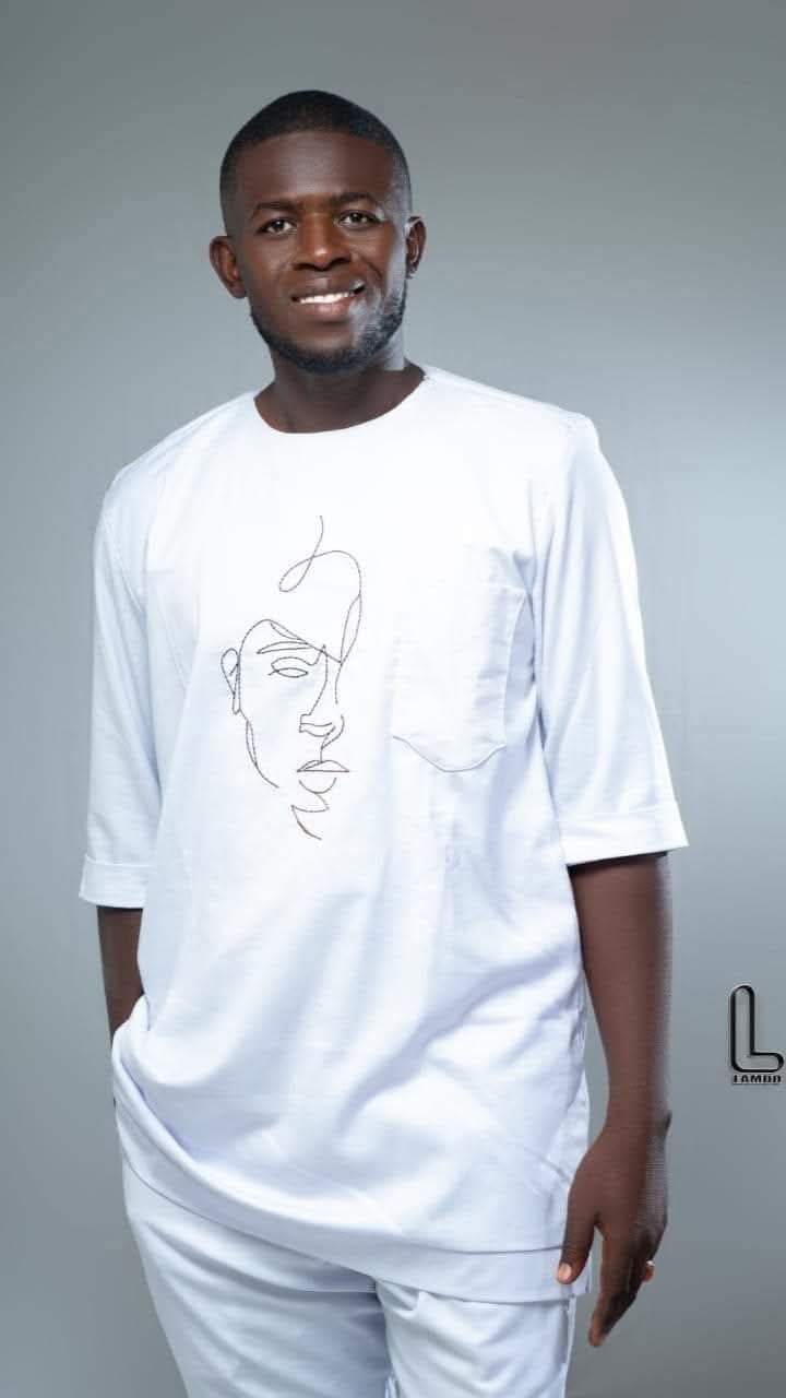 Young man with short hair and beard wearing a white shirt with a line drawing of a face, standing against a gray background.